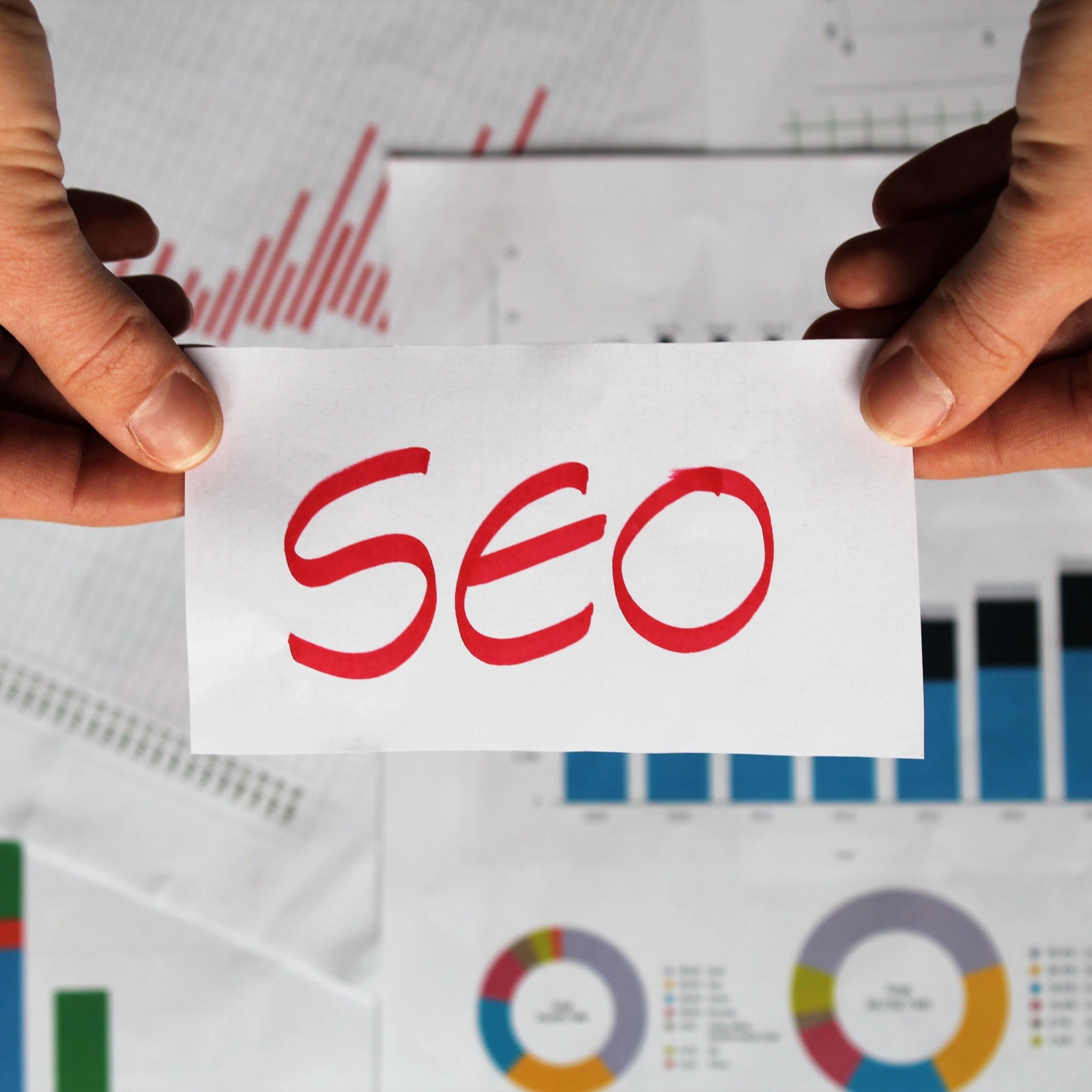 Menschliche Hände halten ein Schild mit der Aufschrift SEO über einem Tisch voller Statistiken