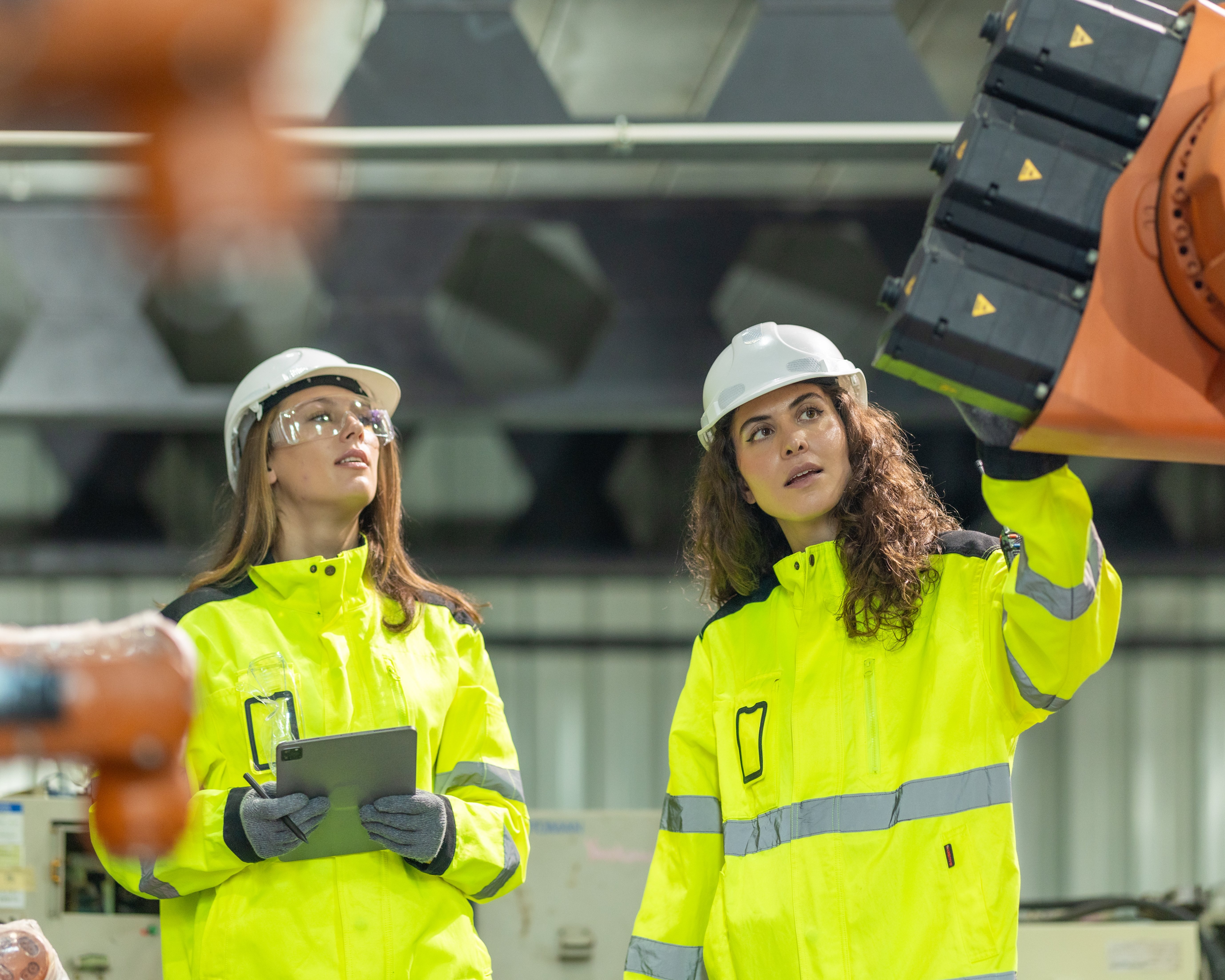 Zwei Ingenieurinnen in Warnschutzkleidung begutachten einen Roboterarm in einer modernen Fabrikhalle